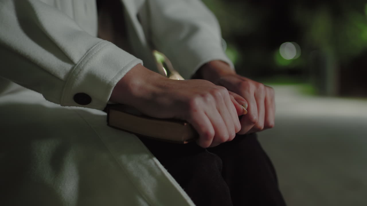 Close up girl in white coat moving book forward with both hands, serene nighttime atmosphere enhanced by distant bokeh lights and soft blur background