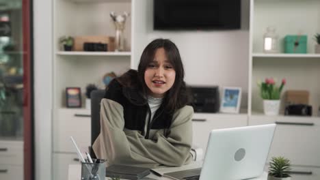 The-portrait-of-a-young-dark-haired-woman,-arrogantly-looking-into-the-camera-and-saying-something,-sits-in-a-bright-room-at-a-table-in-front-of-a-laptop