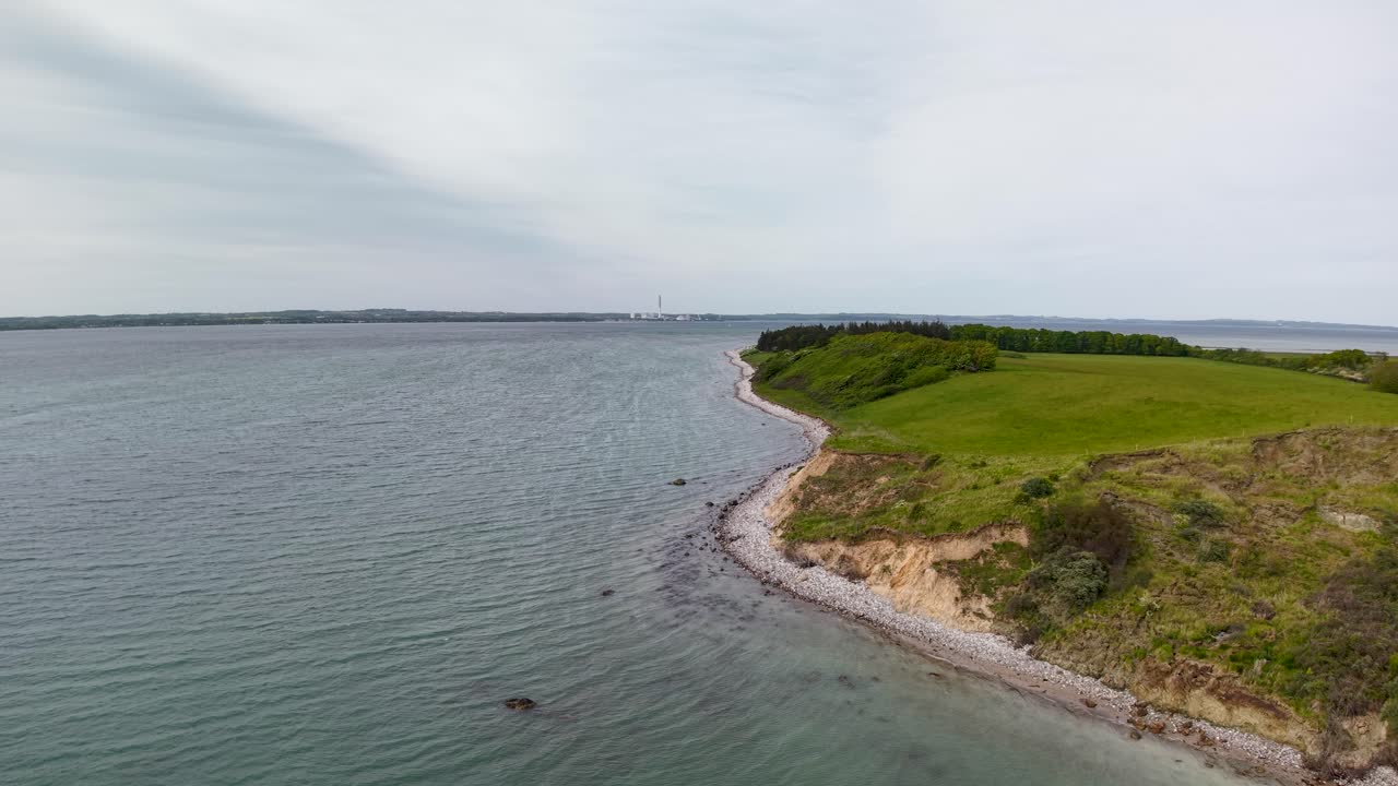 Aerial drone footage of Denmark’s coastal landscape with rocky shoreline, gentle waves, and green vegetation along the water’s edge under cloudy daylight