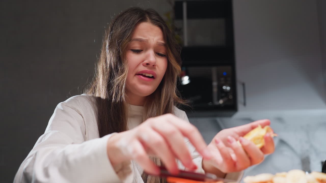 Close view young girl under bright light chewing fruit, arranging slices on table, hands moving, kitchen background, focused expression, shadows, clean counter, fresh snack preparation in progress