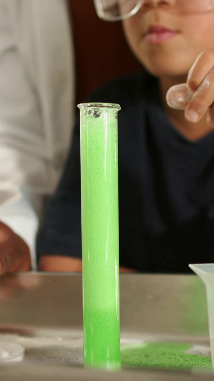 Youth tapping powder onto tube rim causing green foam at lab table, labcoated teacher demonstrating