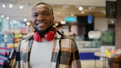 Portrait-of-a-confident-and-happy-man-with-Black-skin-in-a-plaid-shirt-and-red-wireless-headphones-walking-along-the-counters-in-a-modern-grocery-store