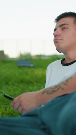 artista de la música disfruta escuchando música al aire libre, acostado de costado con la mano apoyando la cabeza en un campo cubierto de hierba, el fondo presenta un objeto poco claro y una persona caminando en la distancia