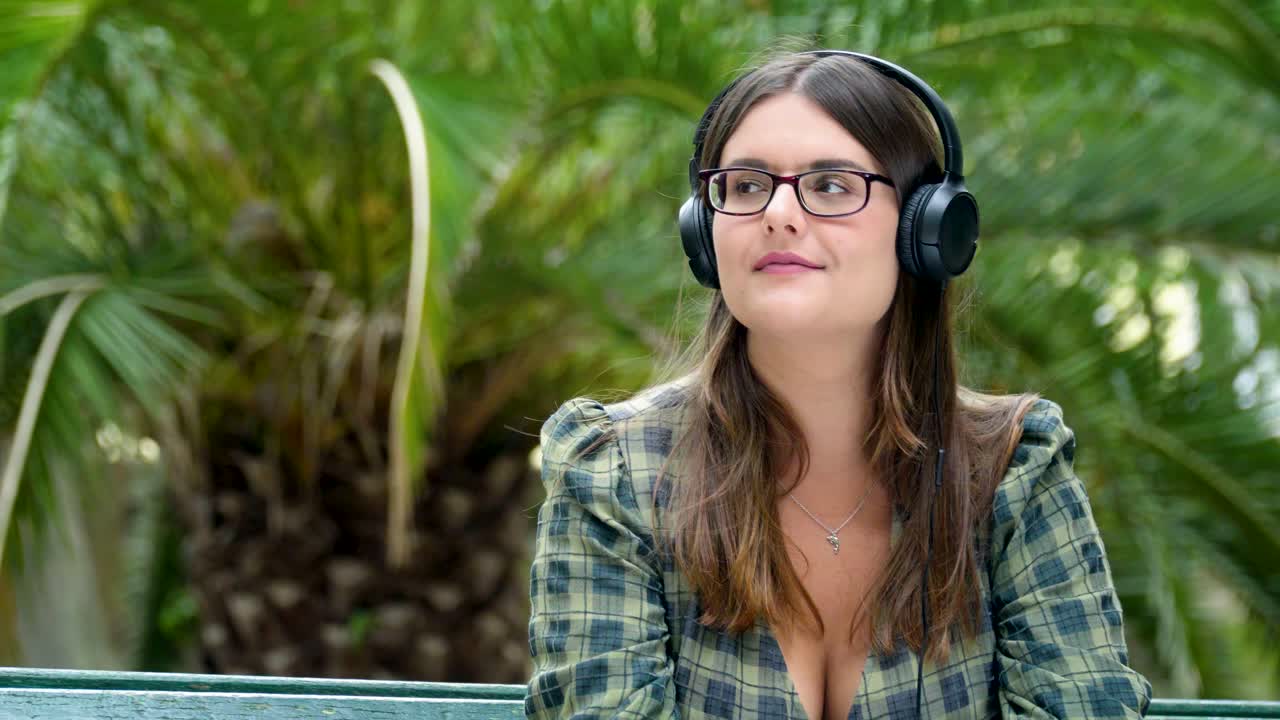 una hermosa chica con los ojos cerrados escuchando música con placer en los auriculares en el parque en un banco con el telón de fondo de una palmera