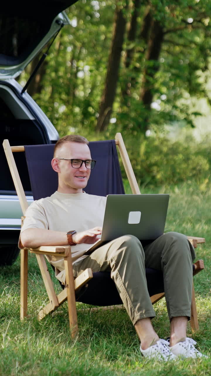 Smiling male freelancer work remote on laptop. Man in glasses is happy to work in nature. Vertical video