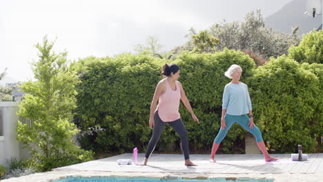 two happy diverse senior women practising yoga by pool in sunny garden, slow motion, copy space