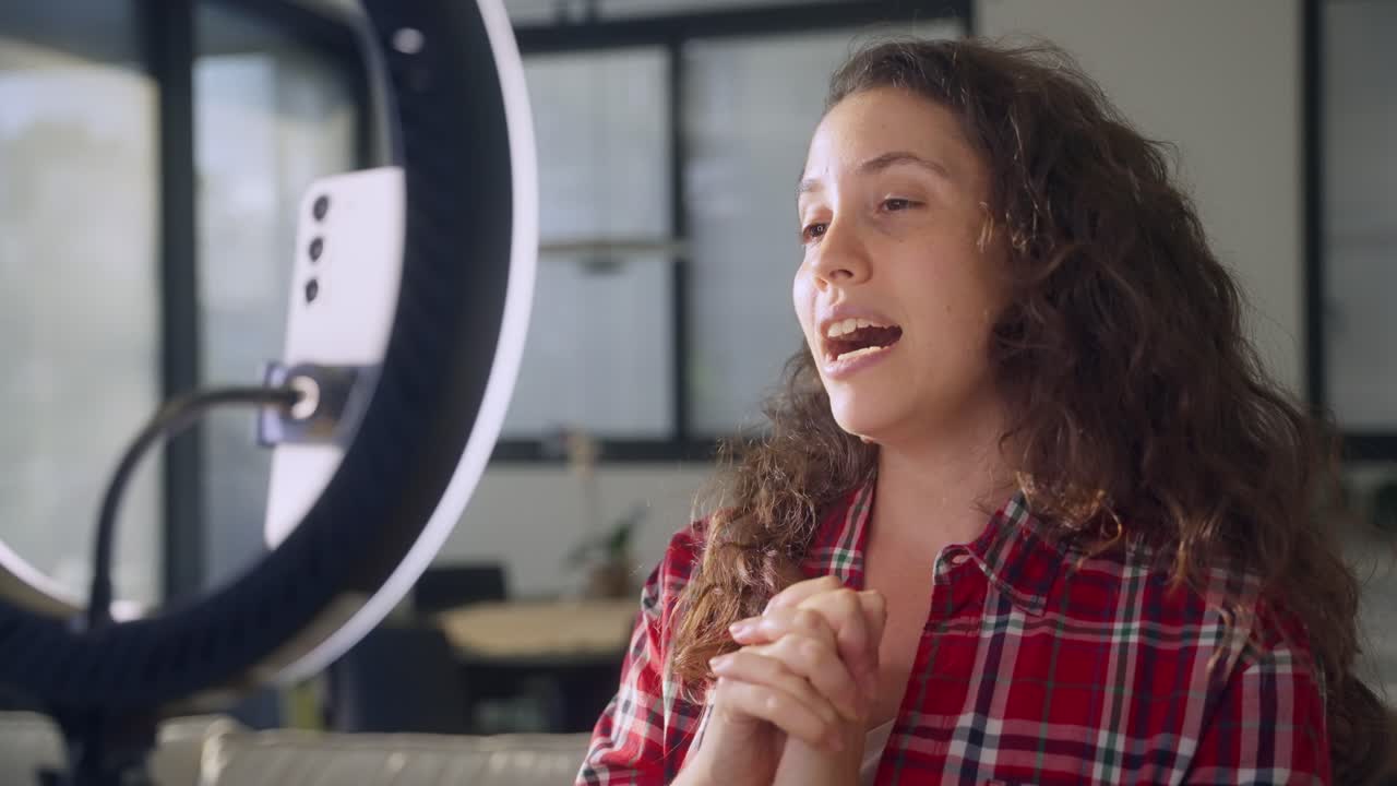 Young social media content creator of influencer using ring light and selfie stand to record herself in home studio or living room. Using mobile phone camera talking to online audience followers fans