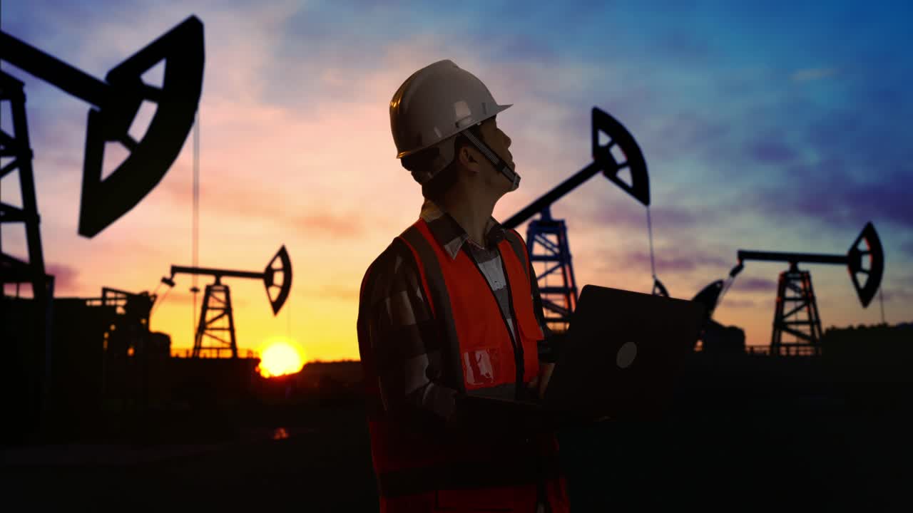vista lateral de un ingeniero masculino asiático con casco de seguridad trabajando en una computadora portátil y mirando a su alrededor mientras está de pie frente a las bombas de aceite, durante la puesta o salida del sol