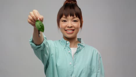 Mujer-Asiática-Feliz-Sosteniendo-La-Llave-Del-Auto-Con-Una-Hoja-Verde.-Concepto-De-Vida-Ecológica,-Medio-Ambiente-Y-Sostenibilidad.-Retrato-De-Una-Joven-Asiática-Feliz-Y-Sonriente-Con-Una-Camisa-Turquesa-Sosteniendo-La-Llave-Del-Auto-Con-Una-Hoja-Verde-Sobre-Fondo-Gris