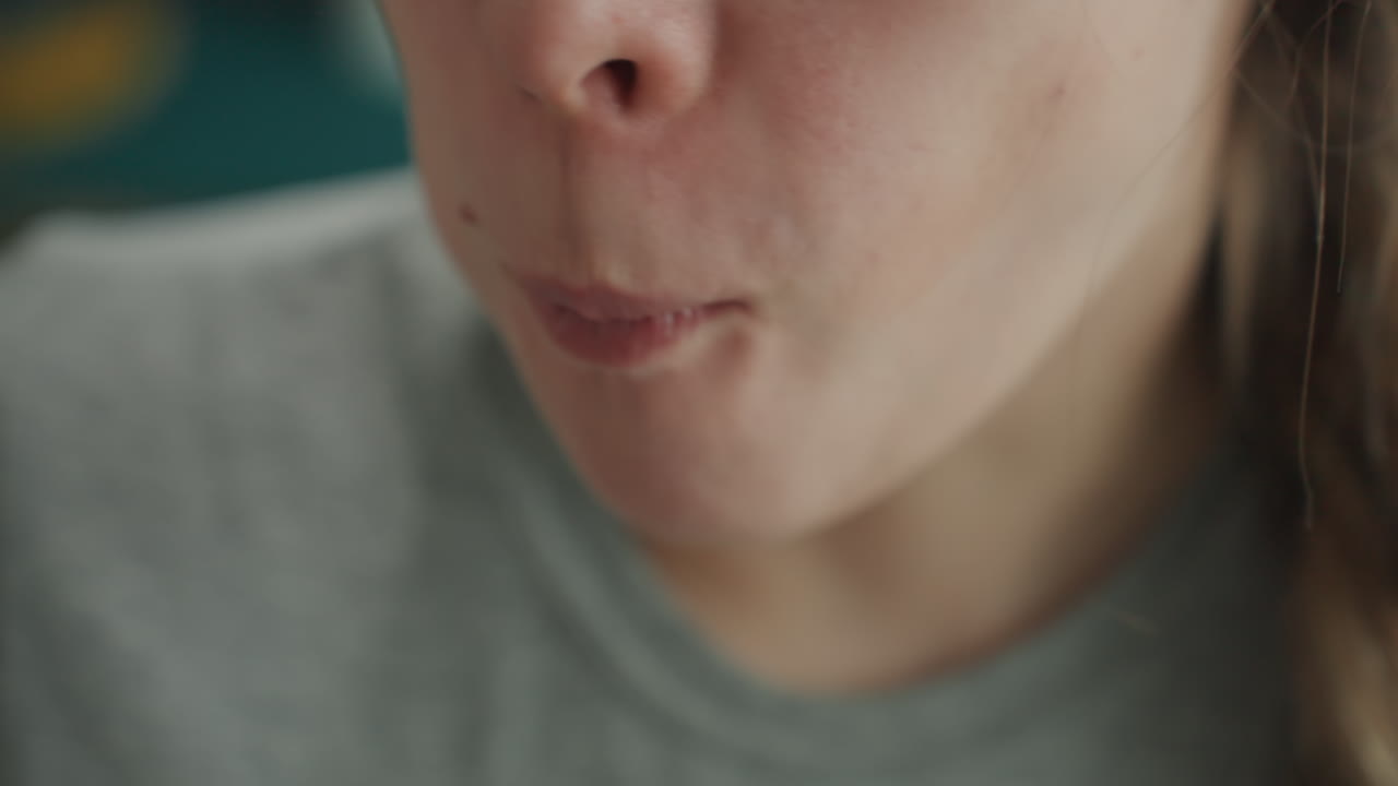 Primo piano di una donna caucasica che assaggia delicatamente il cibo, luce naturale soffusa che illumina labbra e forchetta, masticazione lenta e intenzionale, sottile espressione di contemplazione, maglietta grigia e treccia visibili, sfondo della cucina.