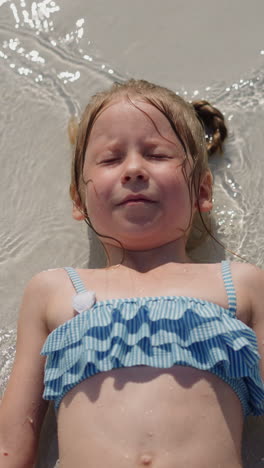 funny little girl in blue swimsuit moves hands lying on sand of surf-line on beach at marine resort on sunny summer day upper close view slow motion