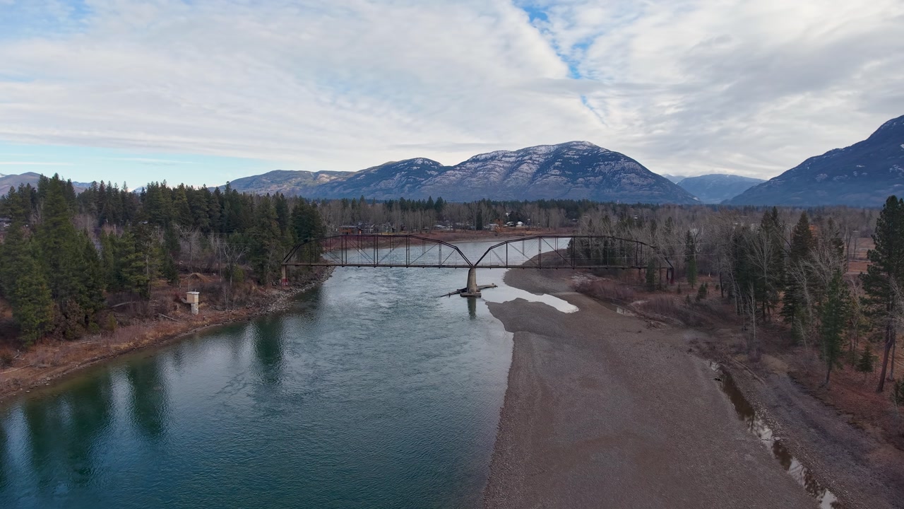Aerial view of a tranquil river flowing under a metal bridge with forested banks and snow-capped mountains rising under a dramatic blue sky.