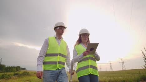 dos ingenieros un hombre y una mujer en cascos con una tableta de ingeniero caminan en el campo con torres de electricidad y discuten la construcción adicional de torres