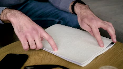 The-Camera-Focuses-On-Man-Hand-Reading-A-Braille-Book-Sitting-On-The-Sofa-At-Home