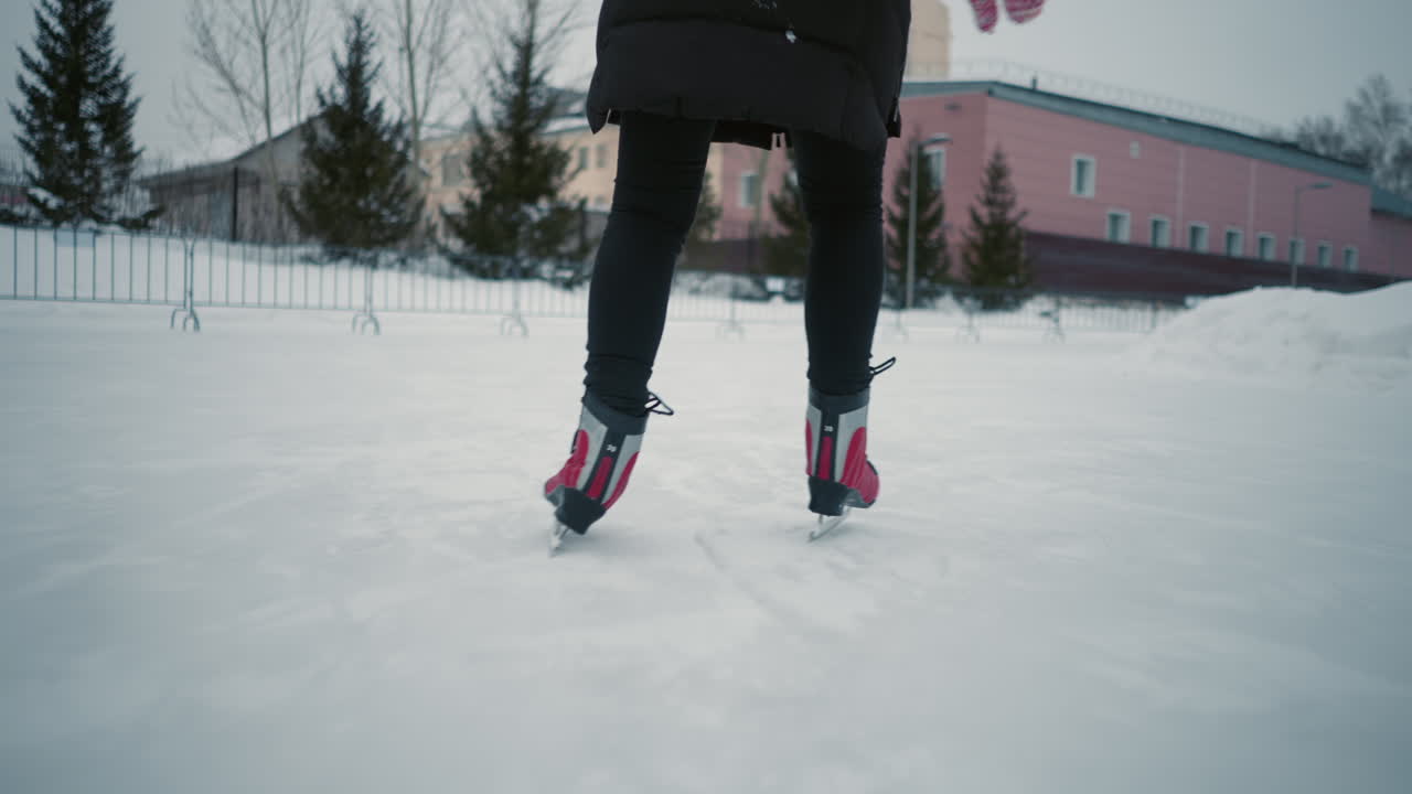 Lady in black winter coat skating on outdoor ice rink, captured from behind focusing on red skates and legs gliding smoothly across frozen surface with snow-covered surroundings and buildings in background