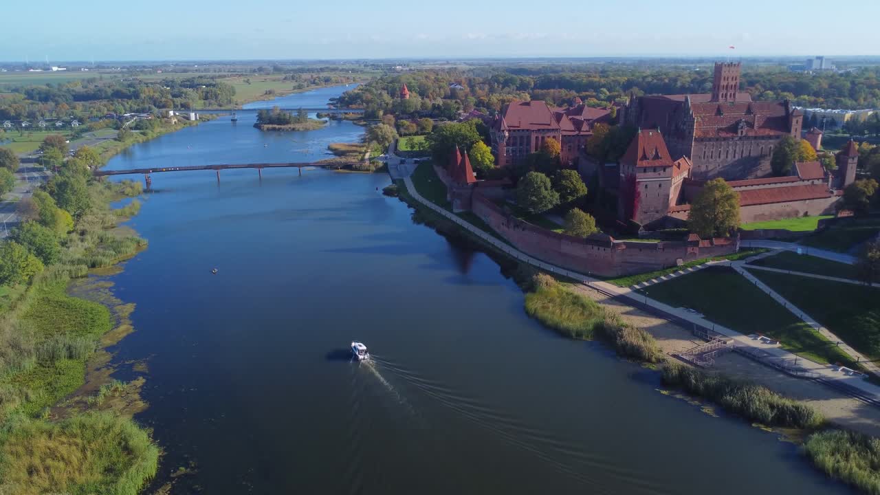Cinematic aerial view of Malbork Castle UNESCO Site with boat travelling along Nogat River in idyllic scene - Malbork, Poland