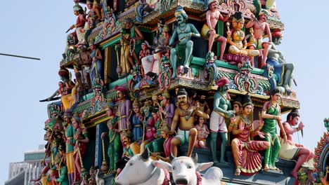 Camera tilts upward along a vibrantly decorated Hindu temple gopuram, revealing detailed statues and carvings in bright daylight, urban background visible