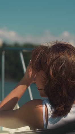 carefree woman with chestnut hair waved by wind rides modern yacht on sunny day. happy tourist enjoys sea cruise on summer vacation. boat sailing