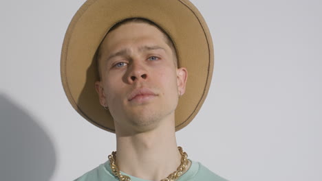 joven modelo posando con un gran sombrero marrón, collar dorado y camiseta verde tocándose la cara y moviendo la cabeza