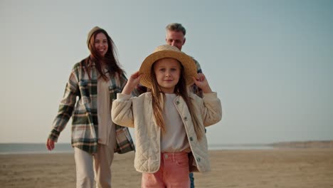 portrait of a happy little blonde girl in a straw hat who looks at the camera and smiles and her parents come up to her from behind and hug her, posing with her on a deserted seashore during a picnic on a hike