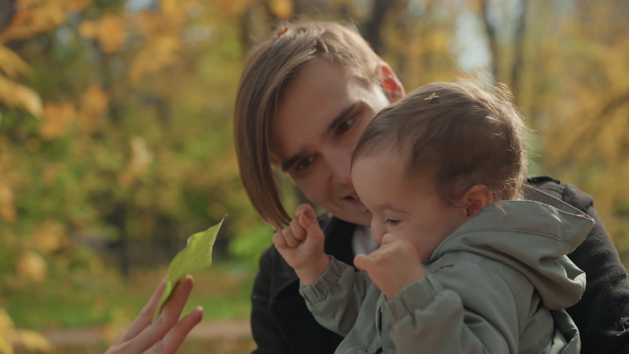 Крупный план: осенняя семья, родитель и малыш исследуют лист. Игривое открытие в парке с золотой листвой, нежное прикосновение и любопытный взгляд. Интимный момент обучения и единения с природой.