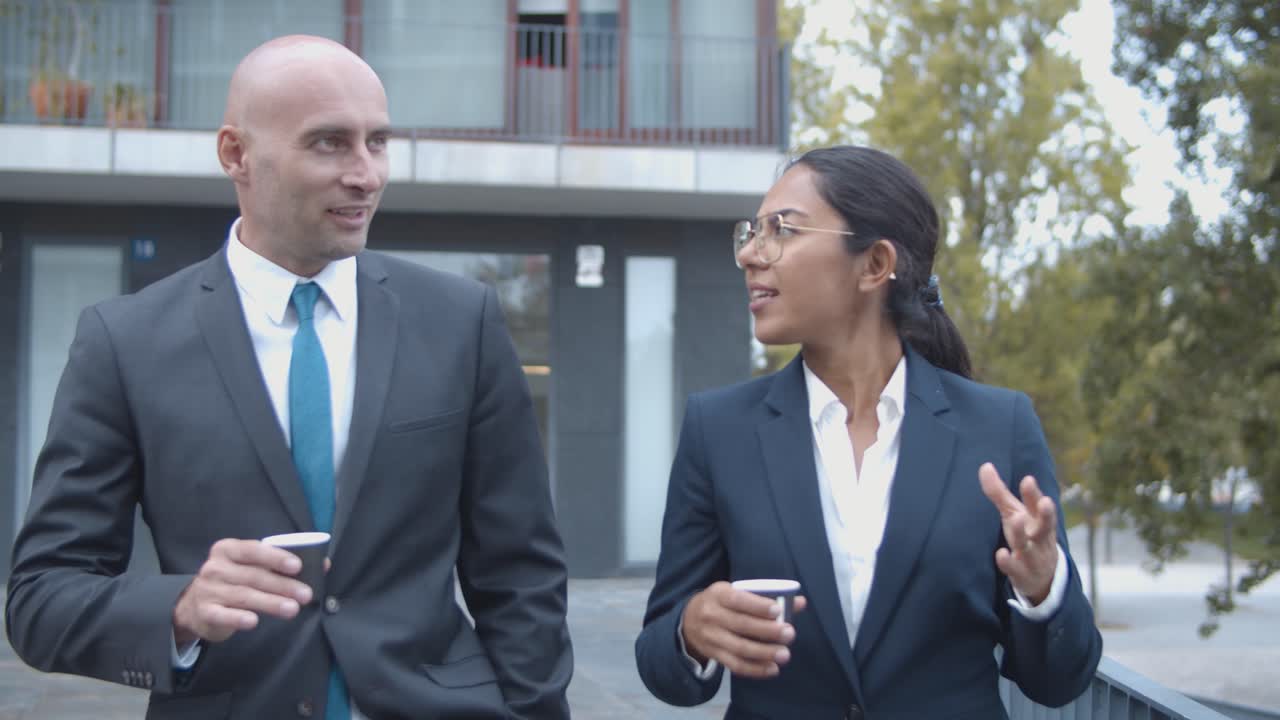 colegas de negocios sonrientes y confiados caminando cerca del edificio de oficinas, bebiendo café y hablando juntos