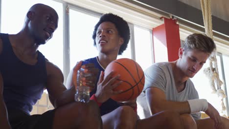 Diverse-male-basketball-team-and-coach-resting-and-drinking-water-after-match