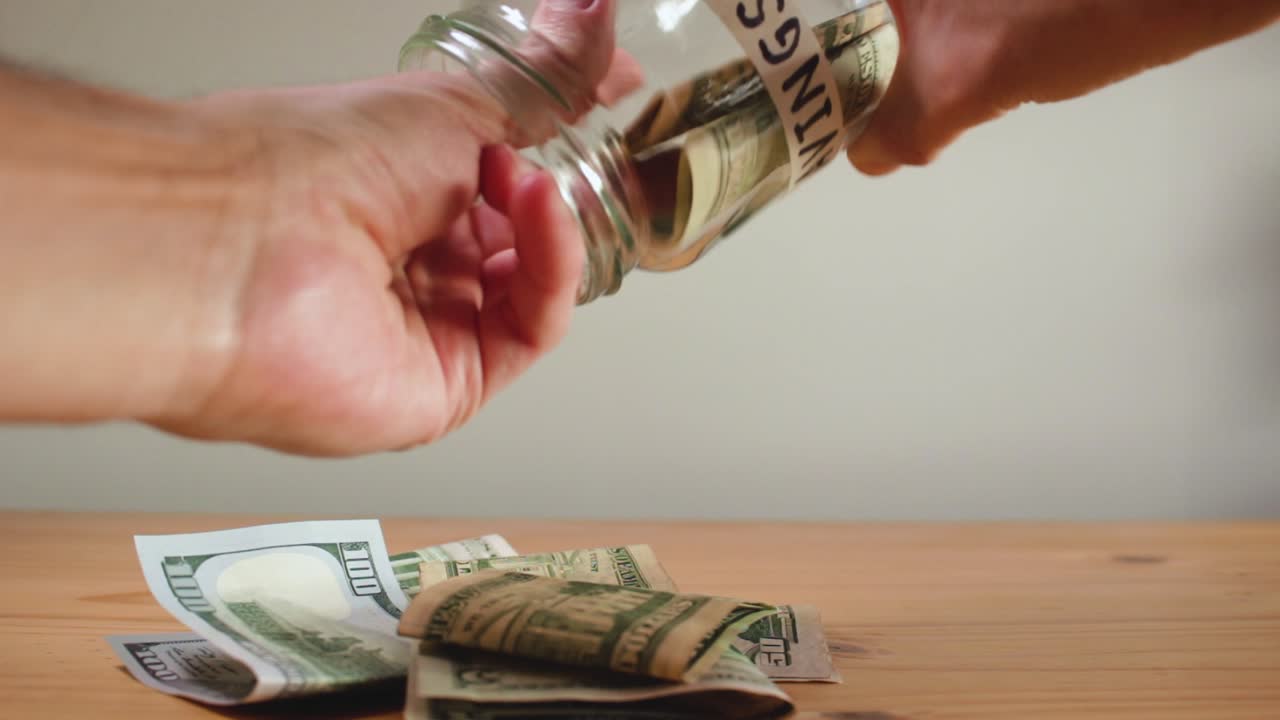 manos del hombre retirando dinero en dólares estadounidenses en efectivo de un frasco de vidrio de "ahorros"