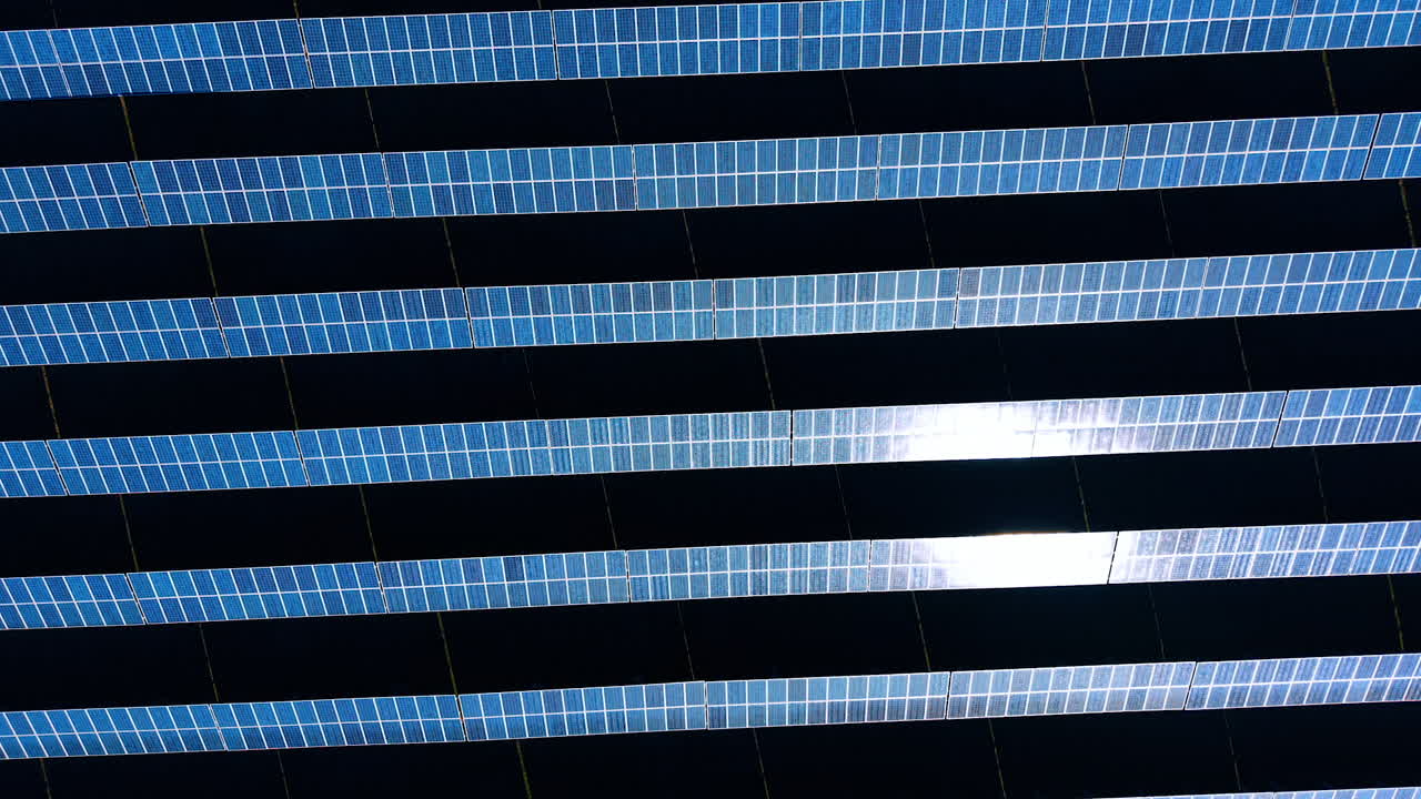 Solar panels arranged in rows on a flat surface under sunlight. Rows of solar panels are set up on a flat surface reflecting light from the sun above