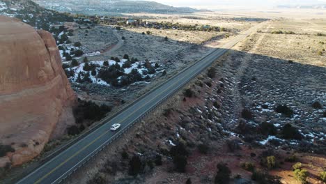 Drone-Siguiendo-Un-Auto-Blanco-En-Moab-Utah-Con-Montañas-Y-Nieve-En-Un-Viaje-Por-Carretera