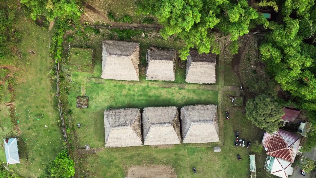 Riprese aeree con drone dall'alto di un complesso di un villaggio tribale indigeno con capanne tradizionali con tetto di paglia, un cortile centrale in erba verde e alberi tropicali.