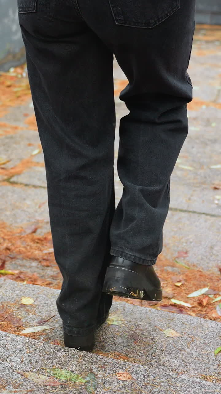Legs of person walking down stone steps during light snowfall, wearing black jeans and black boots, colorful autumn leaves scattered along path, creating peaceful seasonal atmosphere