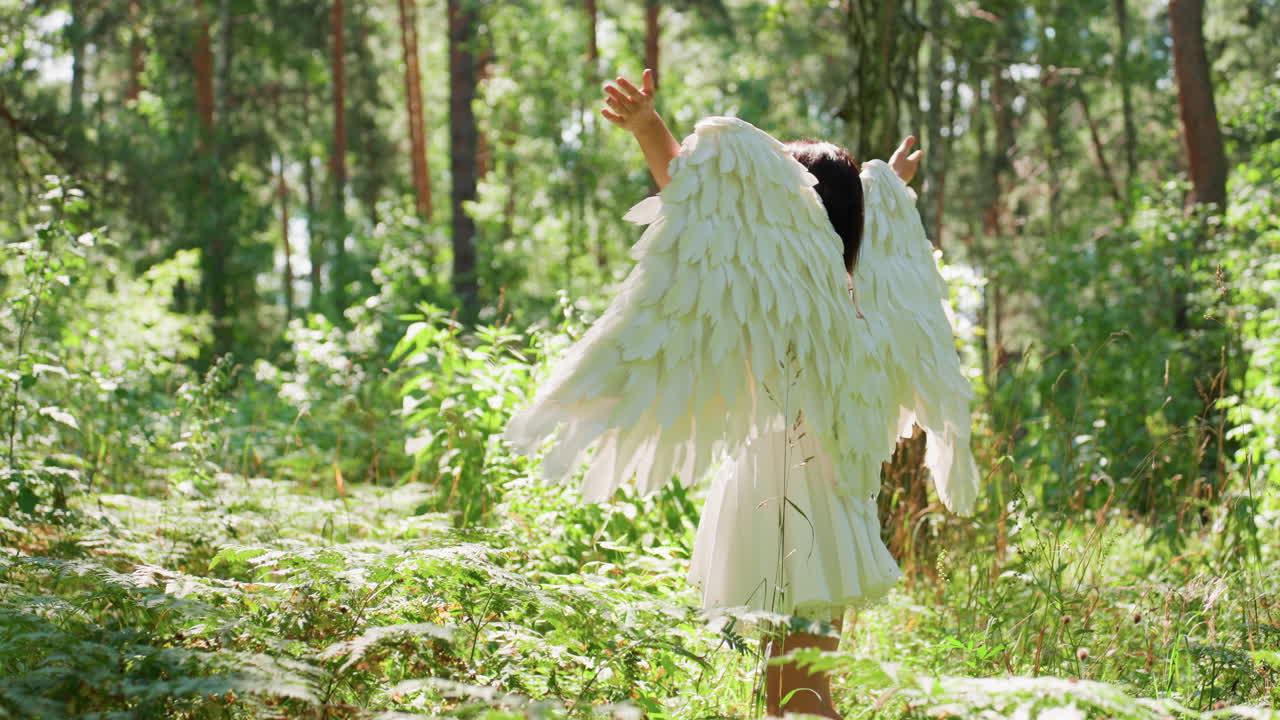 Happy lady wearing angel halloween costume turns around joyfully in lush green forest under warm sunlight, white wings glowing softly while leaves shimmer around