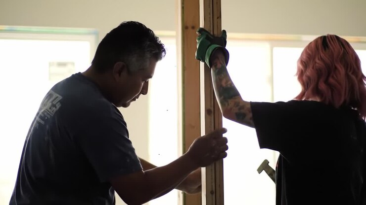 A man and a woman collaborate on a home renovation project in a well-lit indoor area. They are focused on aligning wooden frames while wearing gloves.