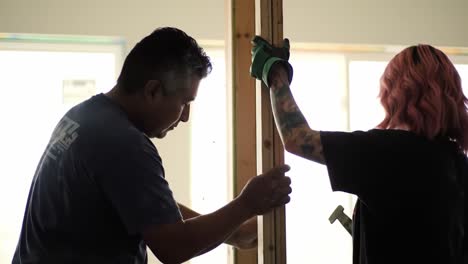 a man and a woman collaborate on a home renovation project in a well-lit indoor area. they are focused on aligning wooden frames while wearing gloves.