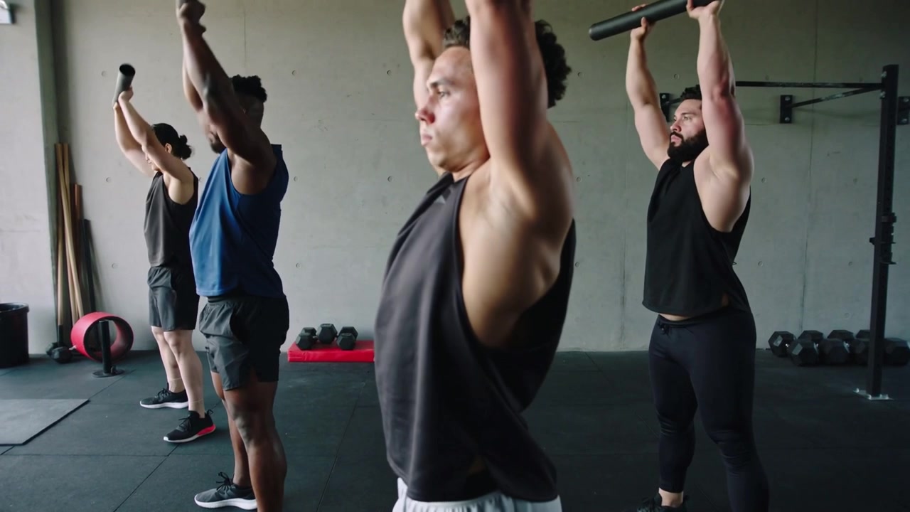 Ein muskulöser Mann im Tanktop hebt auf Kommando gepolsterte Langhanteln über Kopf in einem Fitnessstudio für ein Gruppentraining.
