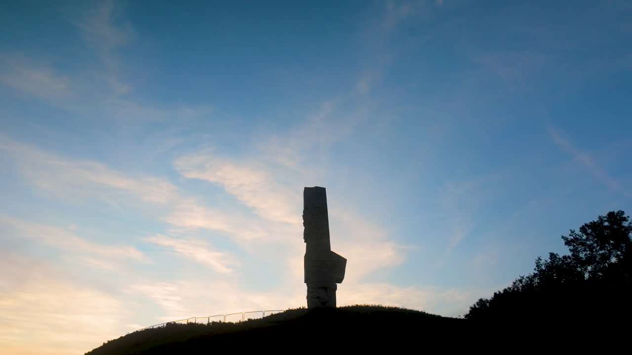 Ultra-wide shot of the Westerplatte monument silhouetted atop its hill against a beautiful, vast blue and orange wispy cloud sunset sky