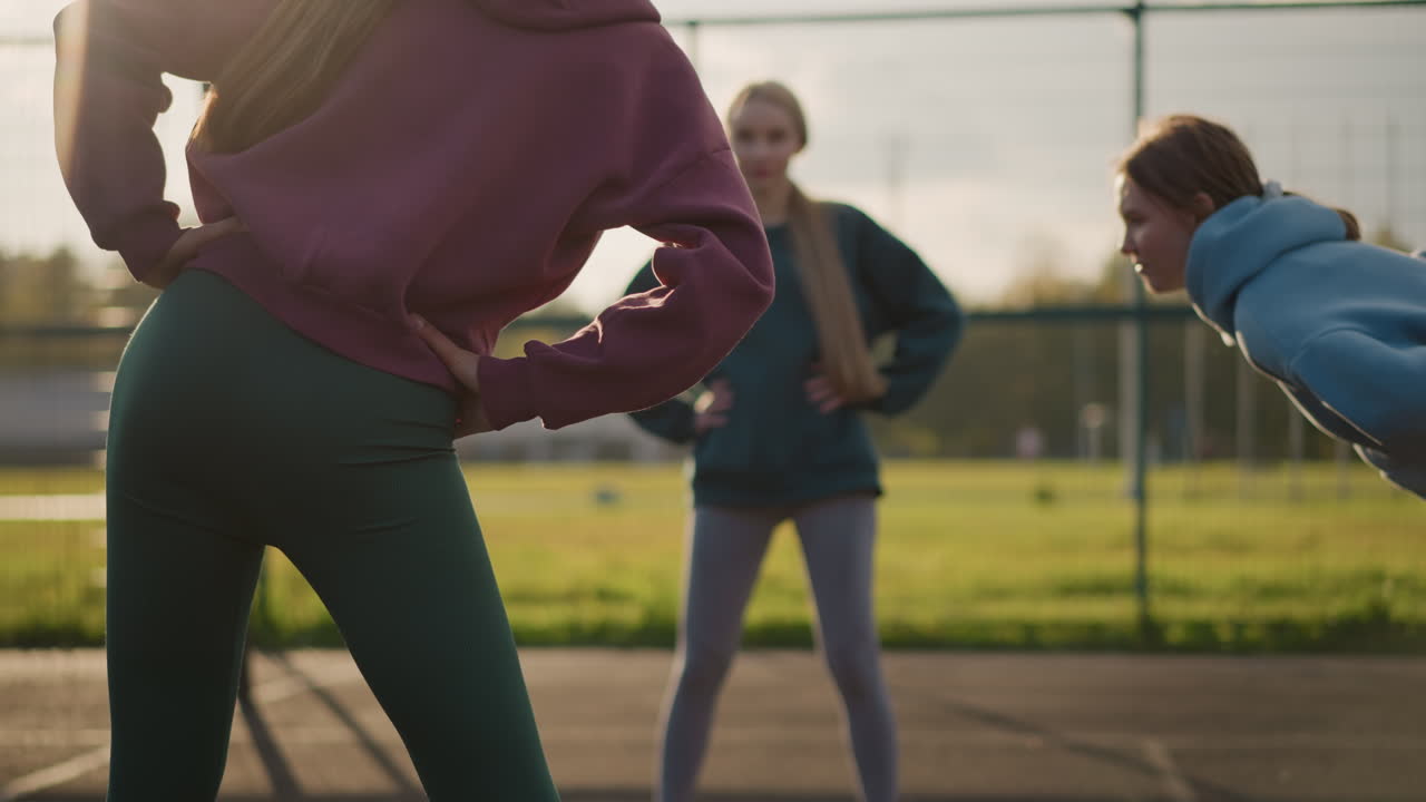 레을 입은 어린 소녀들이 야외 코트에서 피트니스 활동을 하며, 축구장 배경, 빛이 그림자를 던지고, 스트레칭을 수행할 때 역동적인 조명 효과를 만니다.