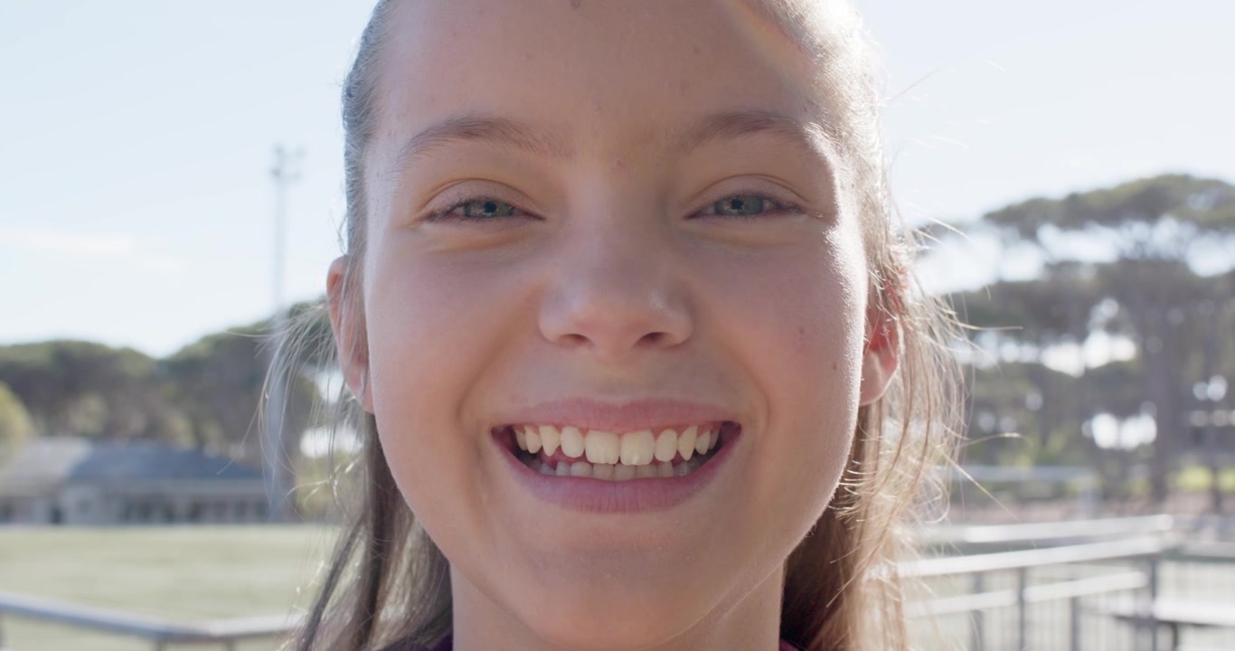 Adolescente respondendo à câmera, entreabrindo os lábios e sorrindo gradualmente na grade do campo esportivo