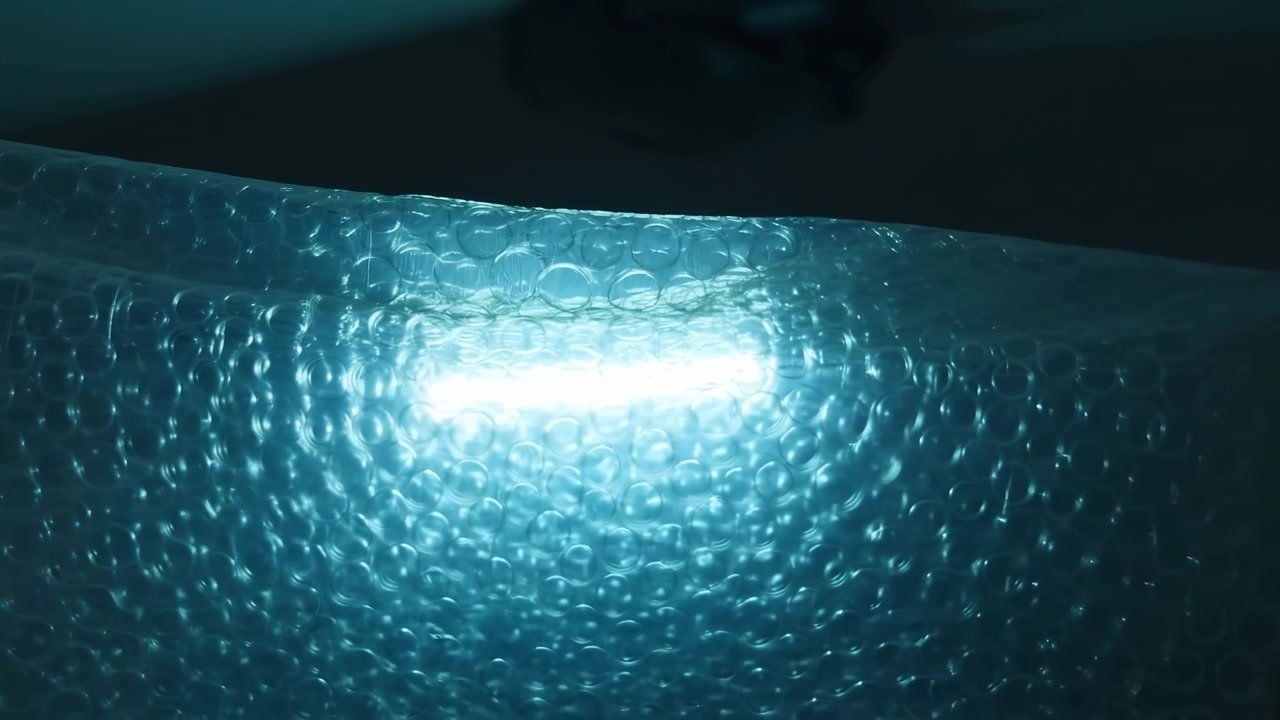 Motion scene of translucent bubble wrap lit from behind by intense cyan glow, producing bright central flare, glossy reflections, curved ridge, repeating circular texture, and deep abstract
