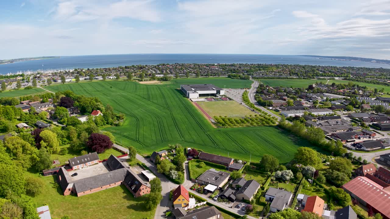 Drone aerial footage showing a Danish town surrounded by green farmland and fields, with a distant coastline and harbor visible under a blue sky