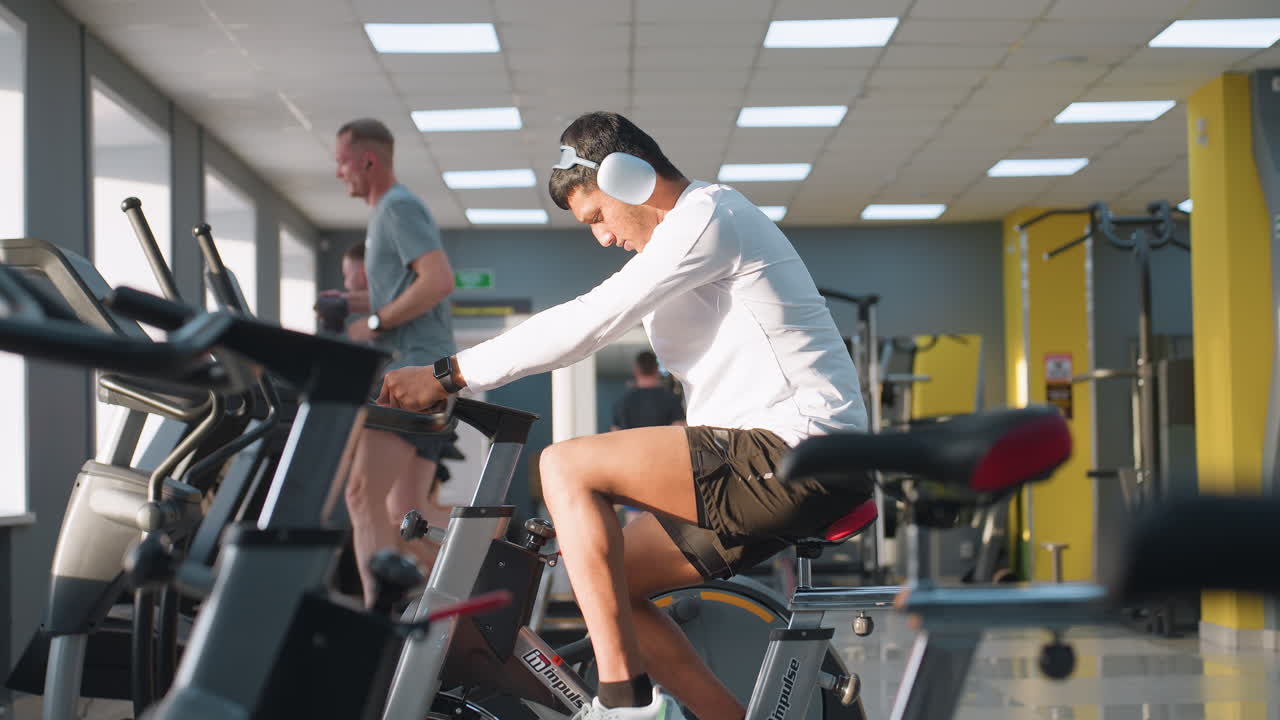 Man in white athletic outfit and wireless headphones cycles vigorously on stationary bike under bright overhead gym lights and natural window light, sweat on skin, with others running on treadmills