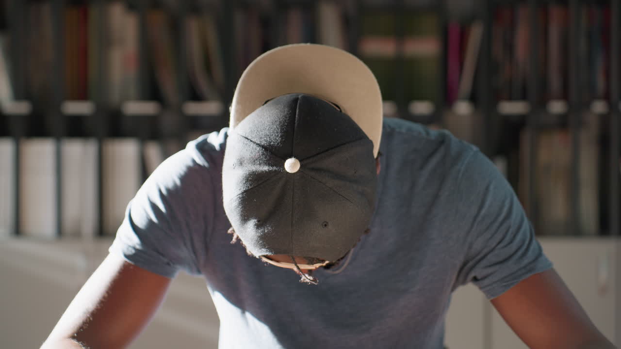 Boy wearing cap sits at library table leaning forward, lifting head slowly from looking down, sunlight highlights profile, stack of books beside him, teal wall and shelves in background