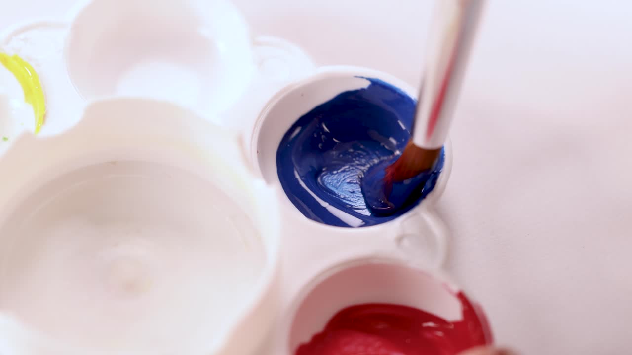 Paintbrush dips into blue paint in white palette, close-up, bright lighting, overhead view