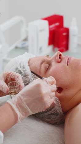 Esteticista-Aplicando-Mascarilla-De-Crema-Hidratante-Exfoliante-En-La-Cara-De-Una-Mujer-Joven-Para-Rehidratar-La-Piel-De-La-Cara