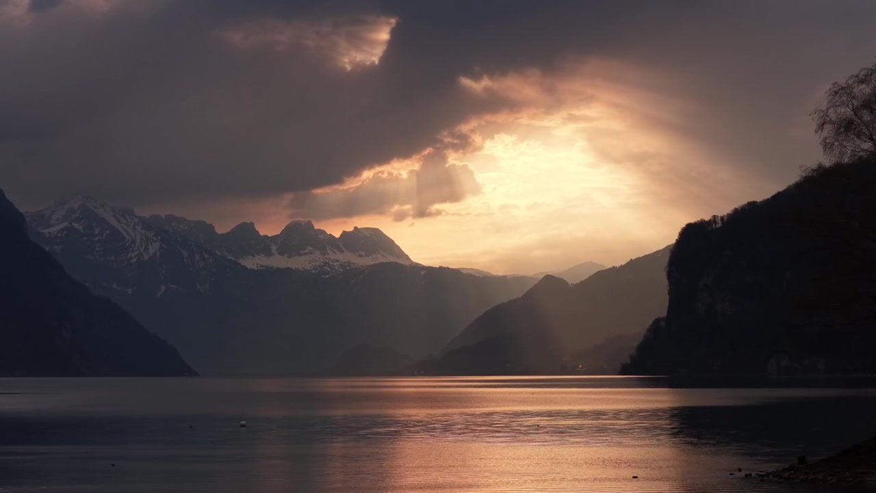 Golden dramatic sunset breaking through storm clouds over alpine lake and snowy mountain peaks Switzerland