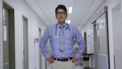 portrait of asian male doctor with hands on hips smiling while standing in the corridor at hospital