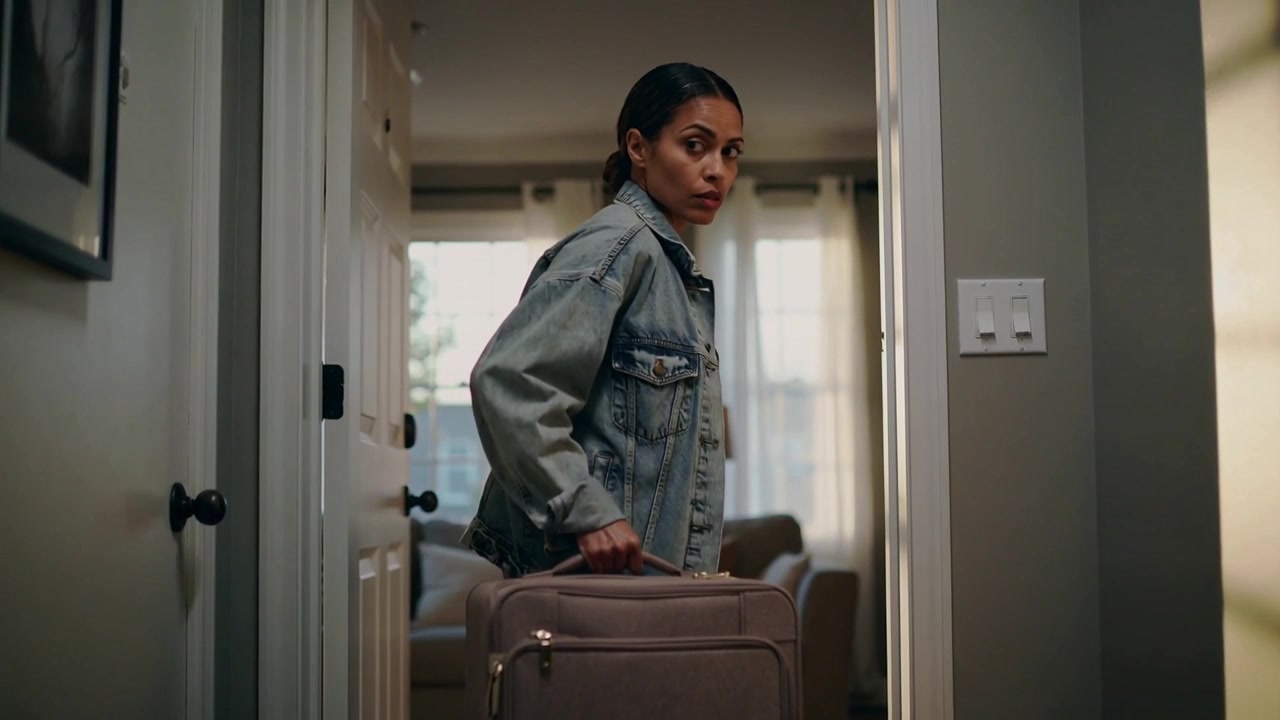 Checking over shoulder, woman in denim jacket moving into living room with suitcase, leaving home