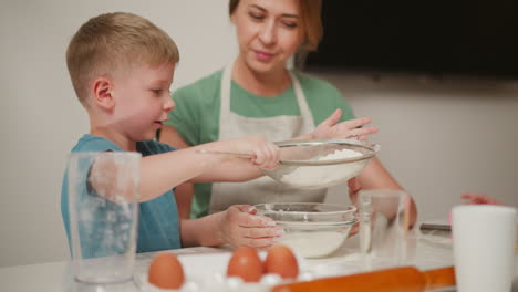 young boy concentrates while sieving flour into glass bowl as woman in apron watches attentively, surrounded by eggs, rolling pin, cups, and whisk on white kitchen table during baking session