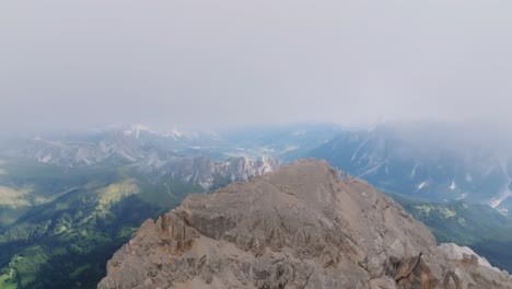 Slow-Aerial-orbit-shot-of-brown-colored-mountain-summit-with-flying-cloudscape-on-top---Beautiful-mountain-range-landscape-in-background-and-rural-valley